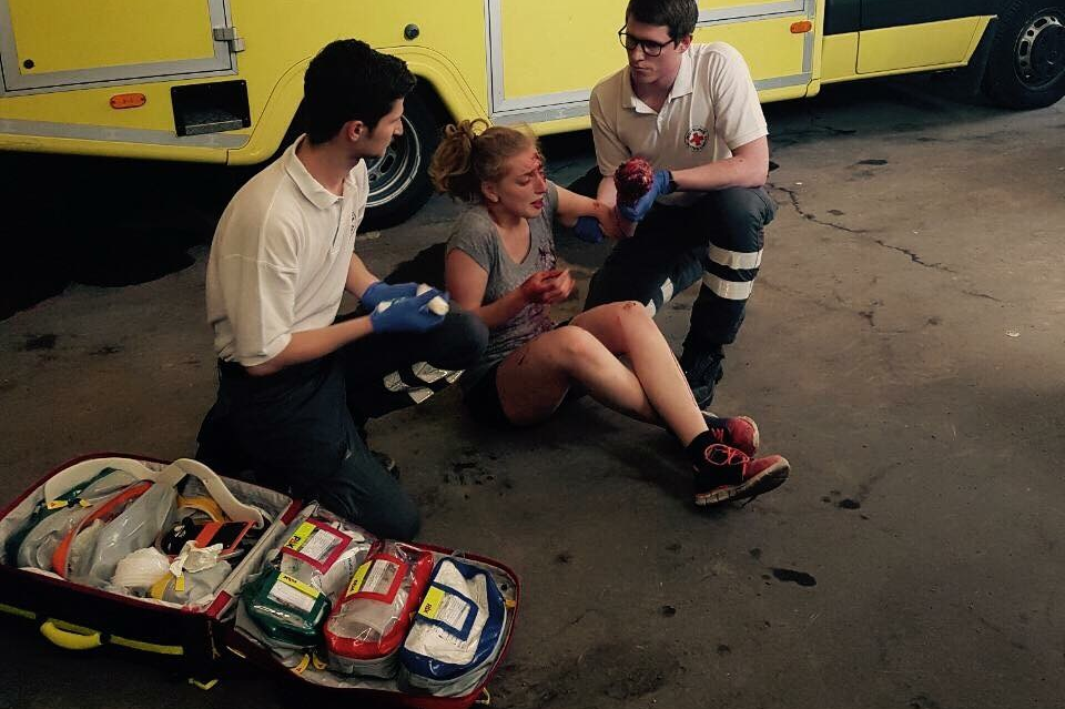 Zwei Helfer knien rechts und links von einer sitzenden Patientin. Es handelt sich um eine Übung. Im Hintergrund ist unser gelber Rettungswagen zu sehen.