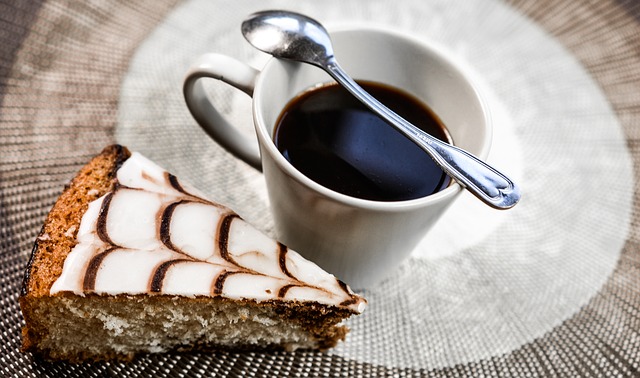 Kaffee und Kuchen Eine Tasse Kaffee. Darauf liegt ein Löffel. Neben der Tasse liegt ein Kuchenstück.