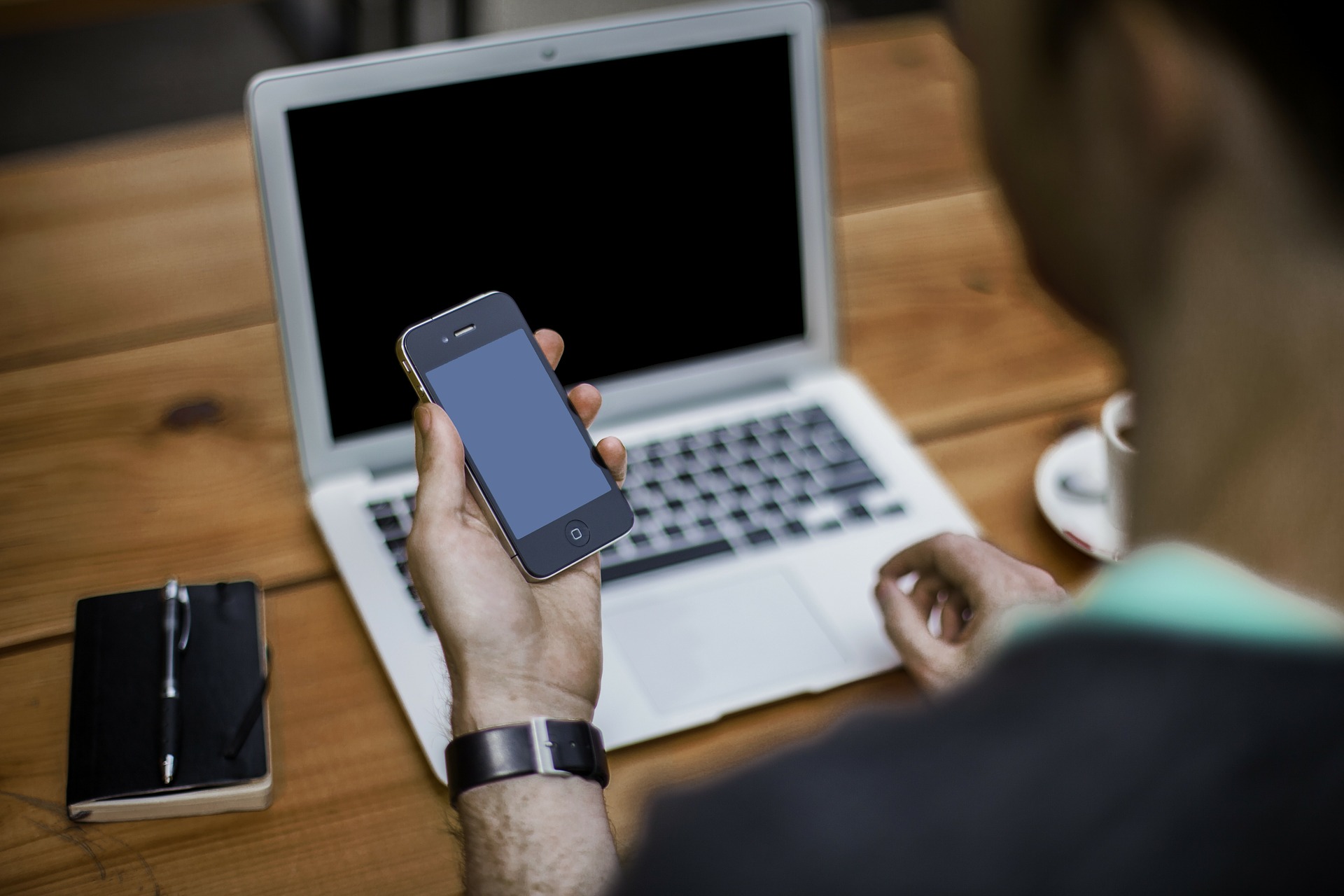 Kontakt Ein Mann sieht auf sein Smartphone und recherchiert gleichzeitig am Notebook